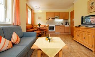 A cozy living room with a blue couch and a wooden table. The kitchen in the background is well-equipped, and there is a television in the corner.