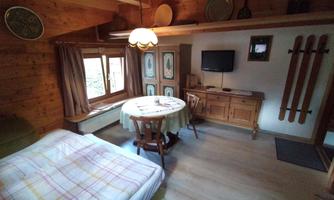 A picture of the living room from another perspective. The spreadable sofa is on the left side, and the view is towards the right side of the room. Next to the window stands a big wooden wardrobe with decorative painting. Next to the wardrobe there is a chest of drawers, and above it there is a TV mounted on the wall. The round table stands between the sofa and the wardrobe. Exit is on the right side of the room.