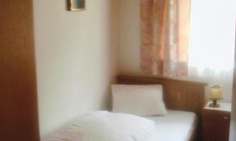 A simple bedroom with a single bed and a bedside table. The window has curtains and there is a small picture on the wall.
