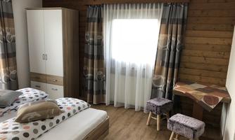 A cozy bedroom with a double bed, a wardrobe, and a small table. The walls are made of wood, and the windows are decorated with curtains.