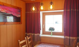 A cozy dining area with wooden paneling and a table. The windows are decorated with red curtains, and a flower is on the windowsill.