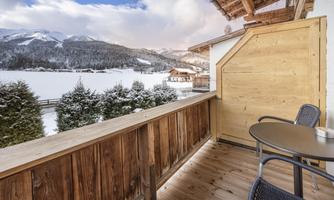 A cozy balcony with a wooden railing and two chairs. In the background, snowy mountains and a clear sky can be seen.