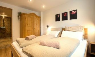 A cozy bedroom with two single beds and a beautiful wooden wardrobe. Pictures of flowers hang on the wall, and the room is brightly lit.