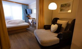 A cozy room with a large bed and a comfortable armchair. There is a television and large windows with curtains that provide plenty of light.