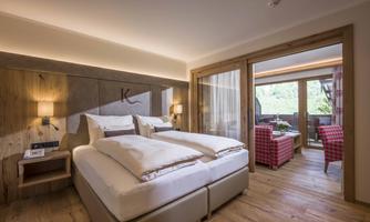 A modern hotel room with a large bed and wooden elements. In the background, there is a sunny balcony with seating.