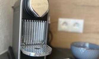 An elegant coffee machine from the brand Krups stands on a work surface. In the background, a power outlet and a bowl are visible.
