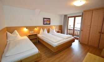 A bright bedroom with two single beds and one double bed. Large windows provide access to a balcony, and the walls are in warm wooden tones.