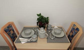 A simple table with two chairs, set with dishes and glasses. In the background, a painting with flowers hangs.