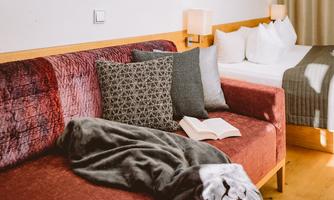 A cozy living room with a red couch, decorated pillows, and a blanket. On the table lies an open book, while in the background there is a bed with white pillows.