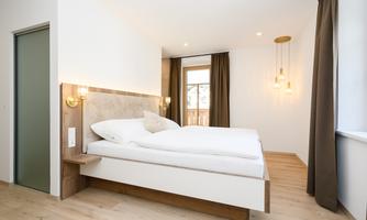 A modern bedroom with a large bed and light wooden floor. The walls are white, and there are elegant windows with curtains.