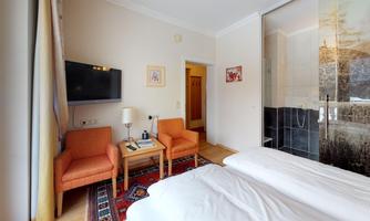 A modern room with two beds, orange armchairs, and a TV on the wall. The open shower is separated by a glass wall.