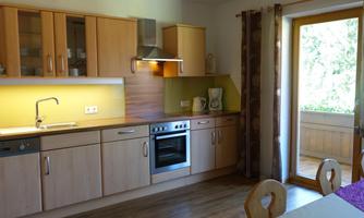 A modern kitchen with wooden cabinets and stainless steel appliances. Behind the kitchen unit, there is access to a balcony with a view of the outdoors.