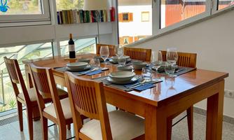 A bright dining area with a wooden table and six chairs. The table is set with dishes and glasses, and large windows let in plenty of light.
