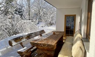 A cozy terrace with a rustic table and benches made of wood. The outdoor area is surrounded by snow and offers a view of wintry trees.