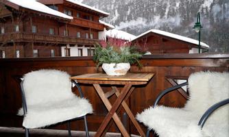 A cozy balcony with two chairs and a small table. In the background, snowy mountains and traditional wooden architecture can be seen.