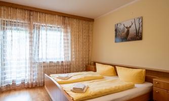 A cozy bedroom with a double bed and yellow linens.  
Large windows with curtains let in plenty of light, and a picture of a roe deer adorns the wall.