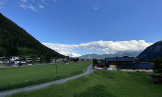 A tranquil landscape with green meadows and small houses. In the background, mountains and a blue sky with few clouds can be seen.