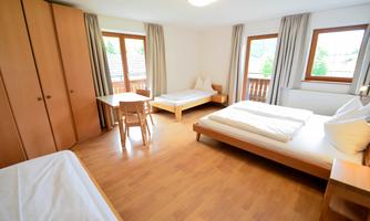 A bright room with two beds, a table, and wooden furniture. Large windows let in plenty of daylight.