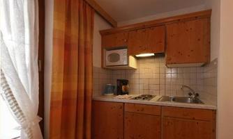 A small kitchen with wooden furniture and a light-colored worktop. There is a window with curtains and a microwave above the stove.
