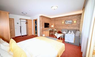 A bright hotel room with a large bed and a cozy seating area. The walls are made of wood, and there is a television as well as a wardrobe.
