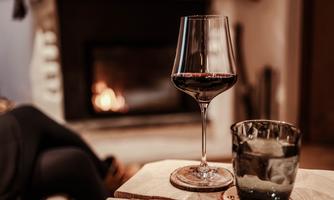 An elegant red wine glass stands on a wooden table next to a glass of water. In the background, a cozy fire is burning in the fireplace.