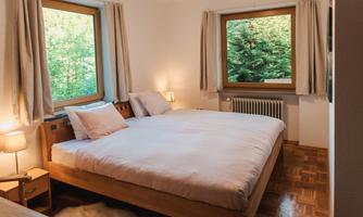A cozy bedroom with a large bed and two windows. The room is bright and friendly, surrounded by natural vegetation.