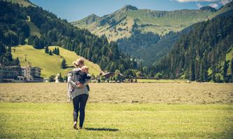 A person with a child on their back walks across a green meadow. In the background, majestic mountains and a blue sky can be seen.