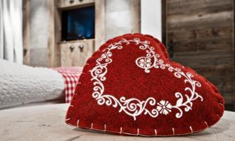 A red heart-shaped pillow with white embroidery is lying on a table. In the background, a cozy, rustic decor can be seen.
