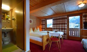 A cozy hotel room with a wooden ceiling and red carpet. There is a double bed, a table, and a view of the mountains through a window.
