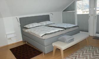 A modern bedroom with a large, comfortable bed and elegant bedding. Next to the bed is a small bench, and the room has large glass elements that let in natural light.