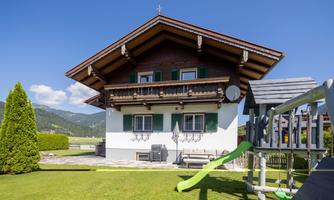 An alpine house with an overhanging roof and green shutters. In the foreground, there is a playground with a slide and a well-maintained lawn.