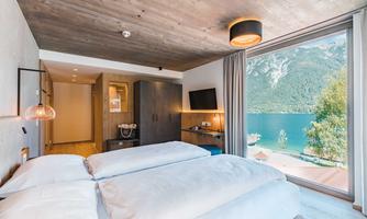 A modern room with two beds and large windows offering a view of the lake and the mountains. The interior is bright and welcoming, with wooden ceilings and stylish furniture.