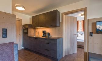 A modern kitchen with dark wood and bright accents. In the background, there is a glimpse of the living area and a bedroom.