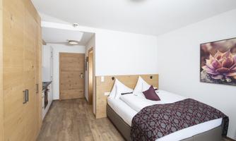 A modern hotel room with a large double bed and wooden details. A picture of a flower hangs on the wall, and there is a small kitchenette.
