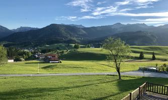 A green landscape with gentle hills and mountains in the background. The sky is clear and there is a quiet village in the distance.
