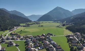 An idyllic landscape with a vast, green valley and mountains in the background. Small houses are scattered near the field.
