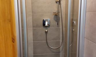 A modern shower with gray tiles and glass doors. The shower is equipped with a handheld showerhead and a shower hose.