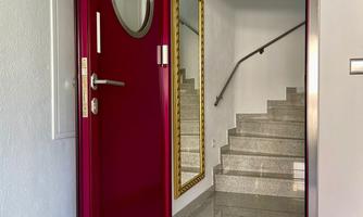 A modern, wine-red front door with a round window. In the background, a staircase and a mirror are visible.
