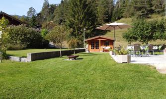 A beautiful garden with a green lawn and a cozy wooden gazebo. In the background, there are trees and there is a dining table with chairs under a sun umbrella.