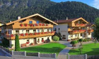 Two cozy buildings with balconies full of flowers surrounded by a well-tended garden. In the background, green hills and a clear sky can be seen.