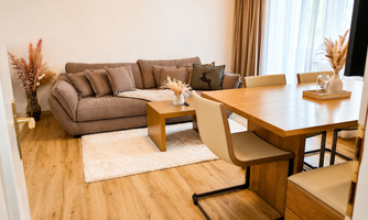 A cozy living space with a beige sofa and a wooden table. The room is bright and friendly, with modern furniture and decorative plants.