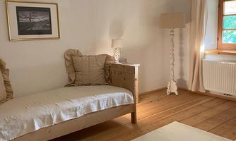 A bright room with a single bed and a table. The room has wooden flooring and a window with curtains.