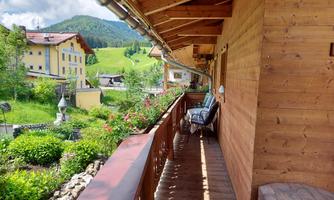 A balcony of a wooden cabin overlooking green meadows and mountains. Blooming plants adorn the balcony.