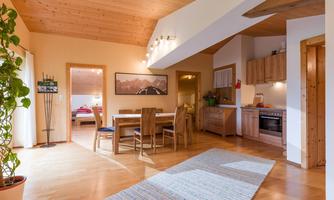 A cozy living space with a wooden ceiling, a dining table, and a kitchenette. The room is bright and friendly with a carpet and plants.