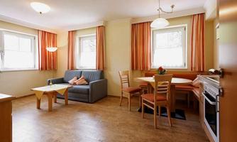 A cozy living room with a couch, a table, and chairs. Brightly decorated with orange curtains and plenty of natural light.
