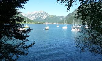 A tranquil lake with several boats and a picturesque mountain landscape in the background. The surroundings are framed by trees and the sky is clear.