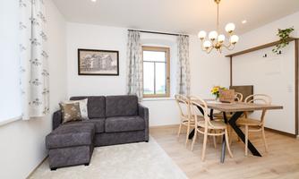 A cozy living room with a gray sofa and a wooden dining table. Large windows let in plenty of light, and the modern decor creates an inviting atmosphere.