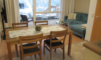 A cozy living space with a dining table and chairs. In the background, there is a sofa and large windows that provide a view of the outside world.