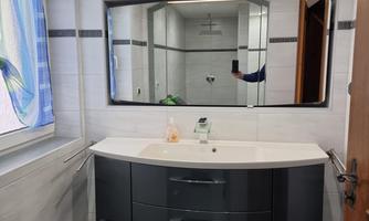 A modern bathroom with an elegant wash area and a large mirror. The walls are light tiled, and the window provides natural light.