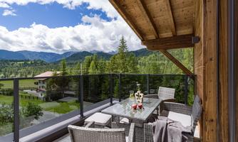 A cozy terrace with a table and chairs made of rattan. In the background, a picturesque mountain landscape stretches out with clouds and trees.